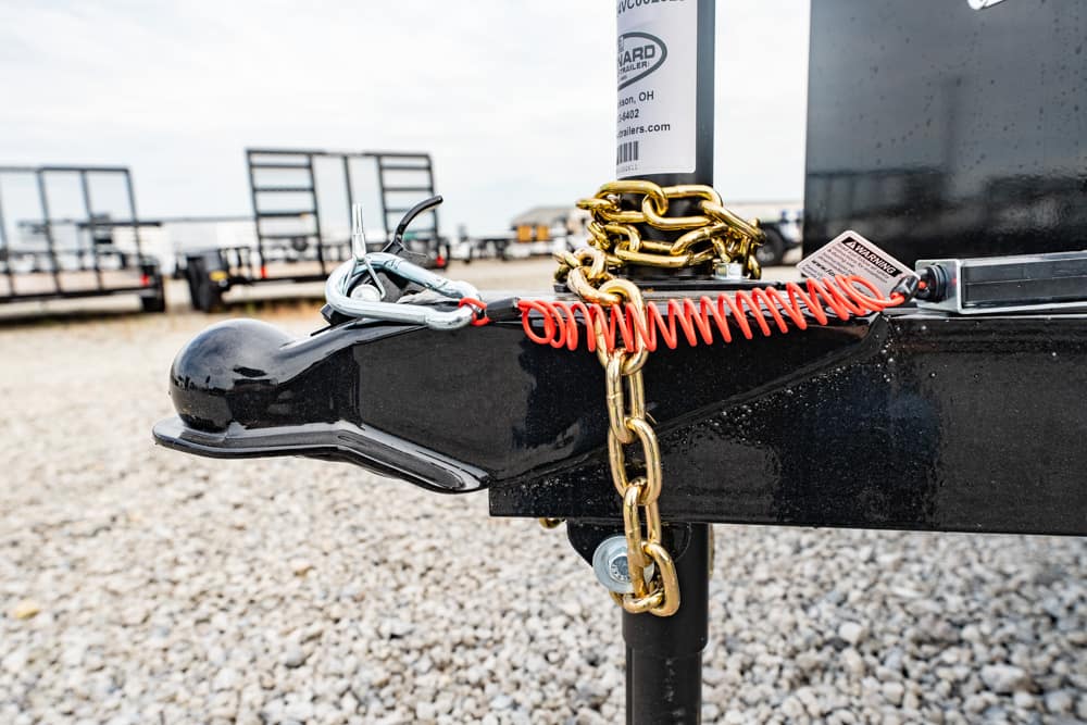 Close-up of a trailer hitch with a gold chain and red locking cable securing the hitch on a gravel lot, with other trailers in the background.
