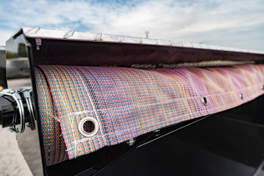 Close-up of a colorful woven tarp with metal grommets rolled into a metal housing on a truck bed, outdoors under a blue sky.