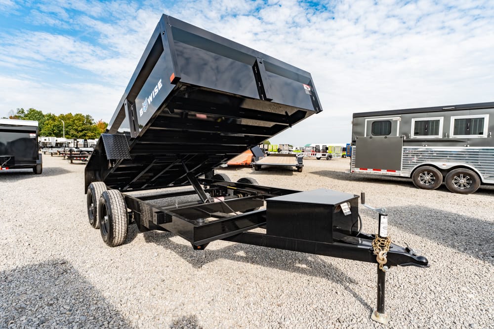 Black dump trailer with its bed raised, parked on a gravel lot with other trailers in the background; front hitch, jack and safety chains visible.