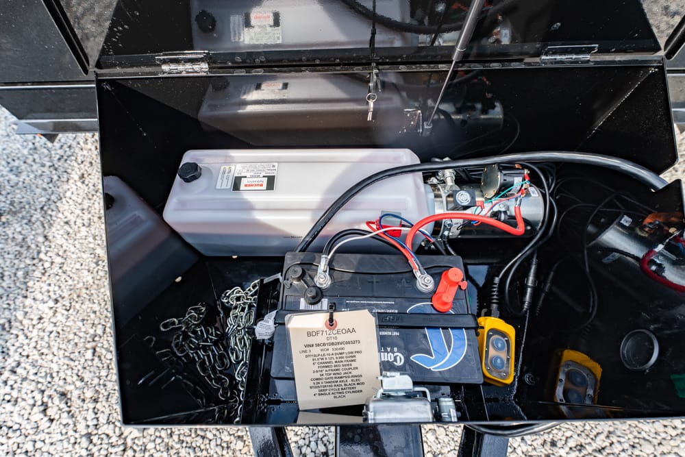 Open trailer battery compartment showing a lead-acid battery with red and black cables, a white plastic reservoir, wiring, and a chain on a gravel surface outside the box.
