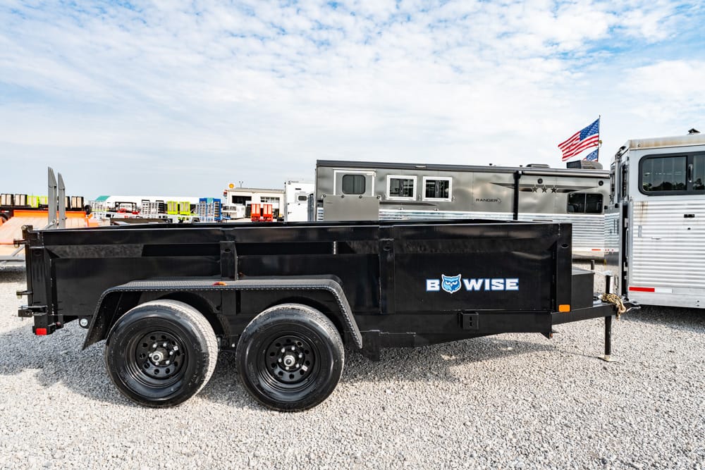 Black dual-axle dump trailer with a raised bed, parked on a gravel lot next to a silver gooseneck trailer; branding reads 'B WISE' on the side with an owl logo; American flag visible in the background.