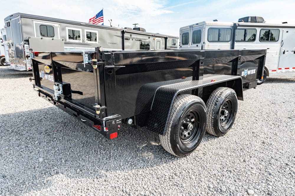 Black tandem-axle utility trailer on a gravel lot with diamond-plate wheels and a tall stake bed; travel trailers are in the background, with an American flag visible above.