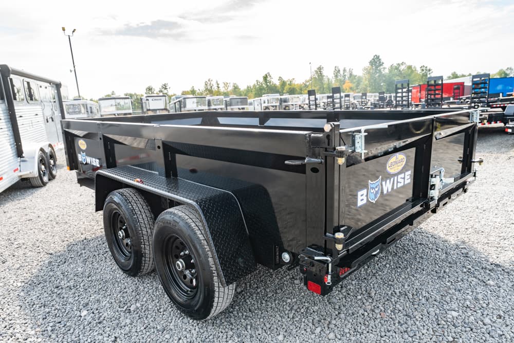 Black dual-axle dump trailer on a gravel lot, with Wise branding and metal fenders, ready for transport.
