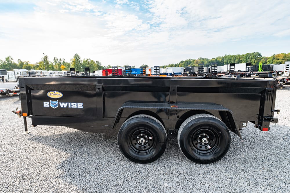 Black professional-grade dump trailer with dual axles on a gravel lot, surrounded by other trailers in a storage yard.