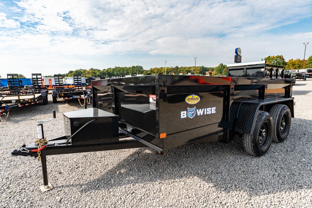 Black tandem-axle utility trailer with a raised bed and dual wheels, parked on a gravel lot with other trailers in the background; brands and decals visible on the side.