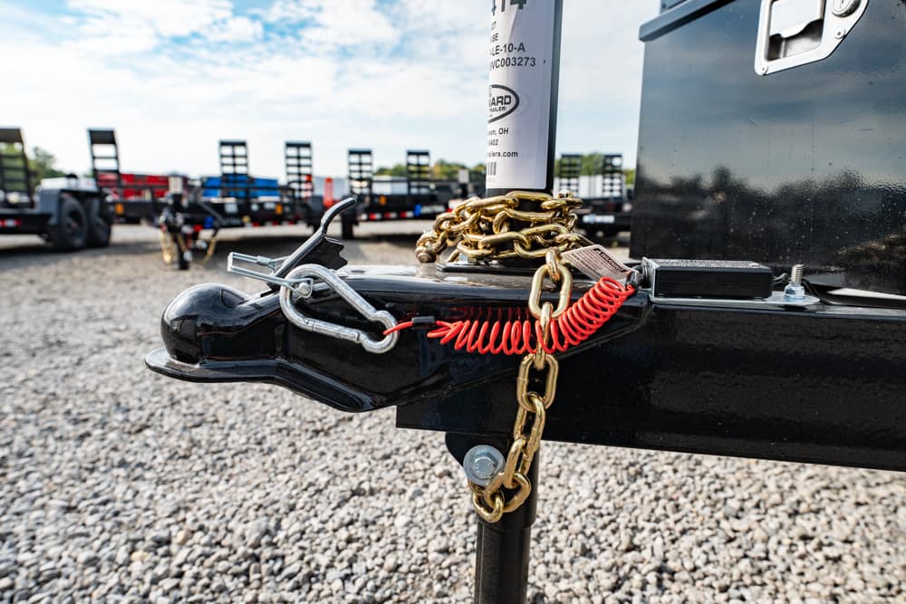 Close-up of a trailer hitch secured with a gold chain and red safety coil on a gravel lot.