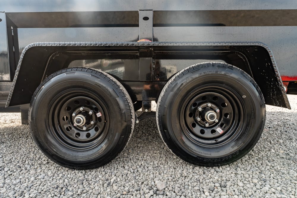 Dual trailer wheels with black steel rims on a gravel surface; tire treads visible under a black fender and utility trailer body.