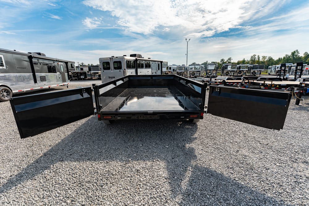 Black flatbed trailer with side doors open, parked on a gravel lot among many trailers under a blue sky.