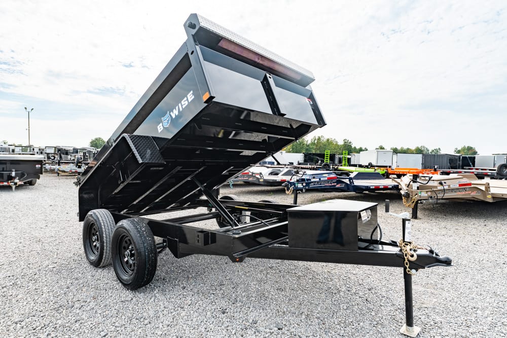 Black double-axle dump trailer with raised bed on a gravel lot, hitch and jack visible.