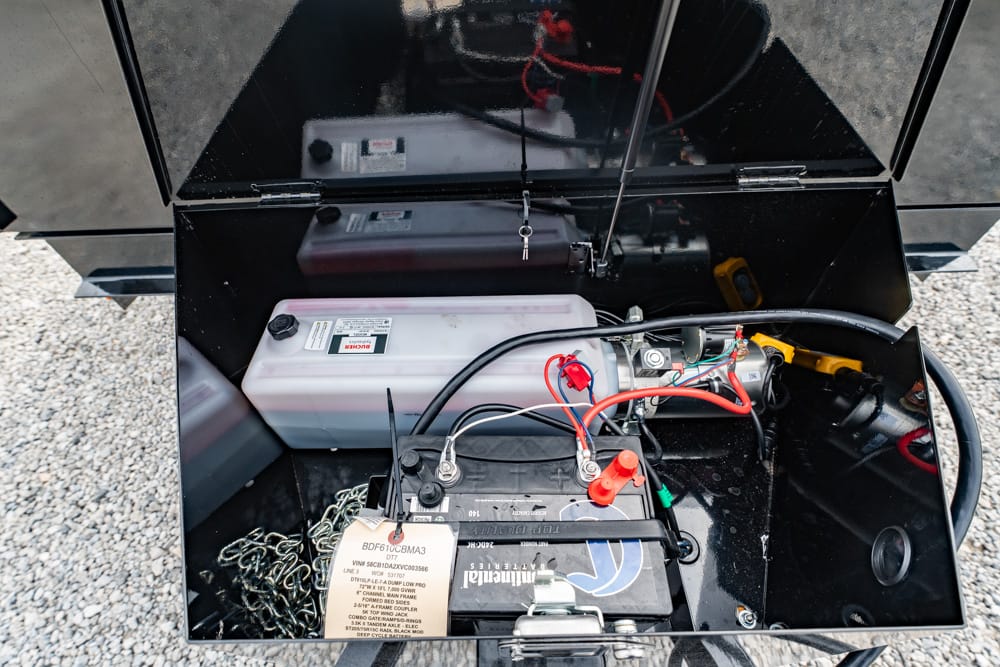 Open metal storage box housing a 12V car battery with red and black cables and various wires inside.