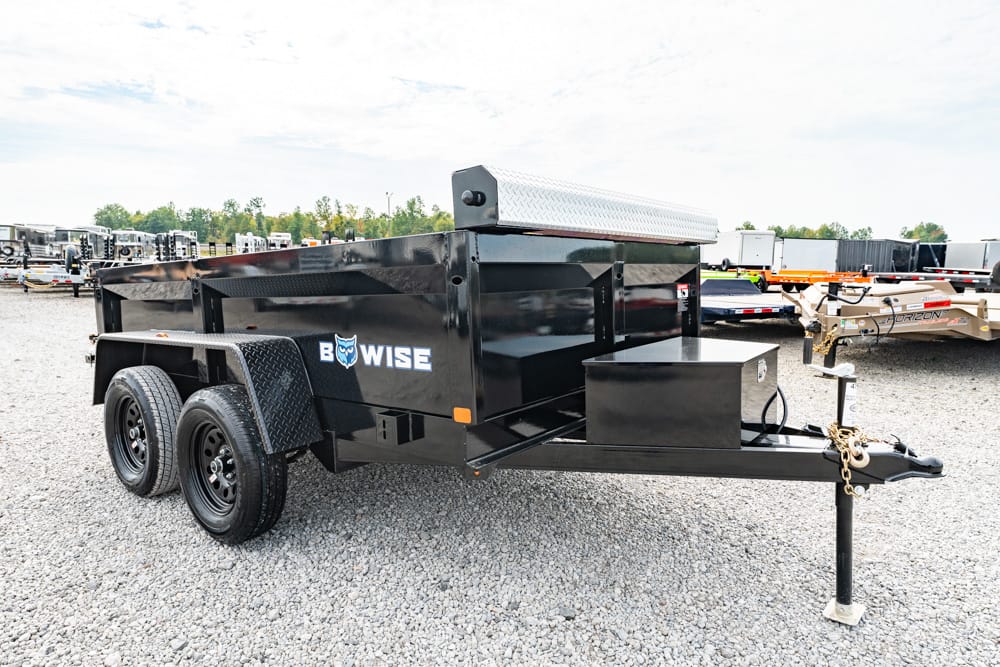 Black Bow-Wise dump trailer with storage toolbox, diamond-plate fenders, and dual-axle wheels on a gravel lot.
