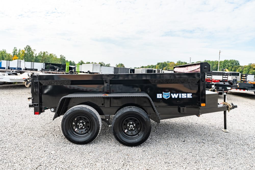 Side view of a black B-WISE dump trailer with dual axles on a gravel lot, ready for hauling equipment or debris.