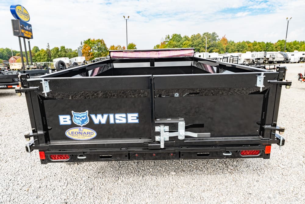 Rear view of a black Bow Wise dump trailer with a split tailgate and metal latches, parked on a gravel lot with other trailers in the background.