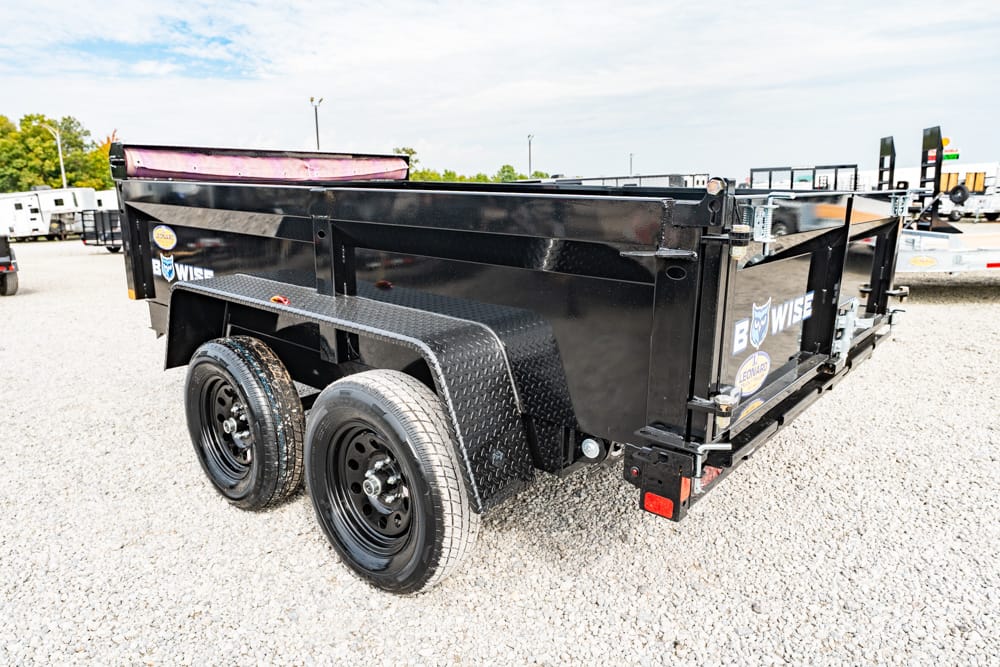 Black dual-axle dump trailer on a gravel lot, showing diamond-plate fenders and side branding.”