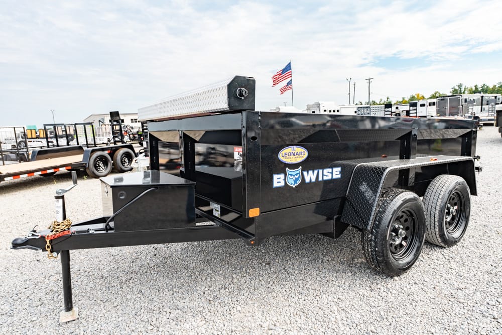 Black tandem-axle dump trailer with a diamond-plate fender, showing the B-WISE logo on a sunny gravel lot.