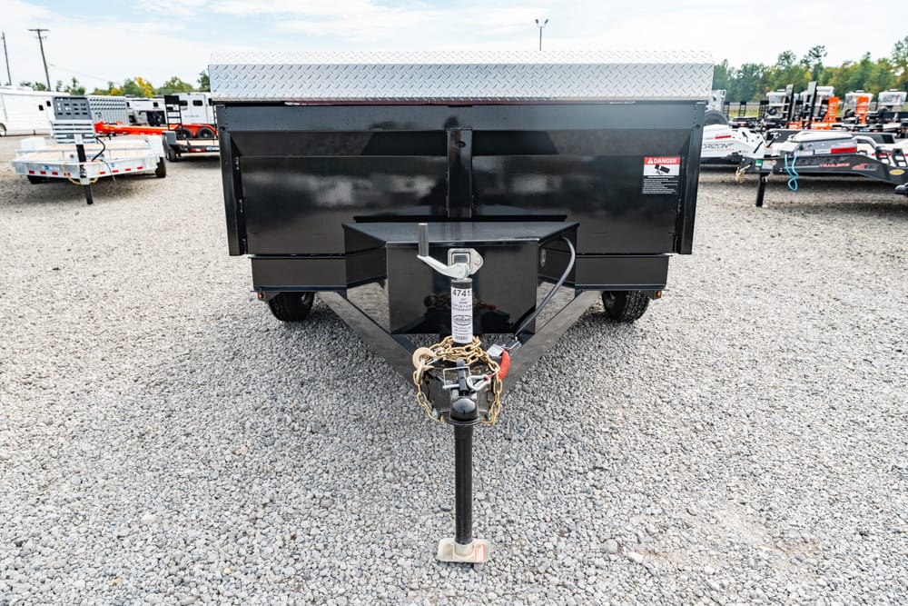 Black dump trailer on a gravel lot, front hitch and jack stand visible with safety chains and pin, surrounded by other trailers in the background.