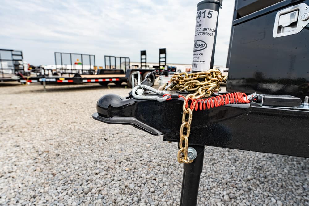 Close-up of a trailer hitch with a gold chain and red safety wire secured on a black frame in a gravel lot with trailers in the background.