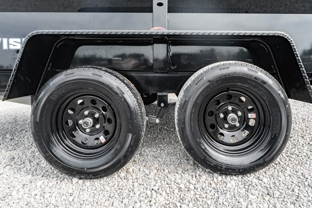 Two black steel trailer wheels with tires on a gravel surface, mounted under a black fendered trailer.