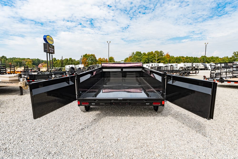 Black flatbed trailer with its side gates swung open, parked on a gravel lot among other trailers under a blue, cloudy sky.