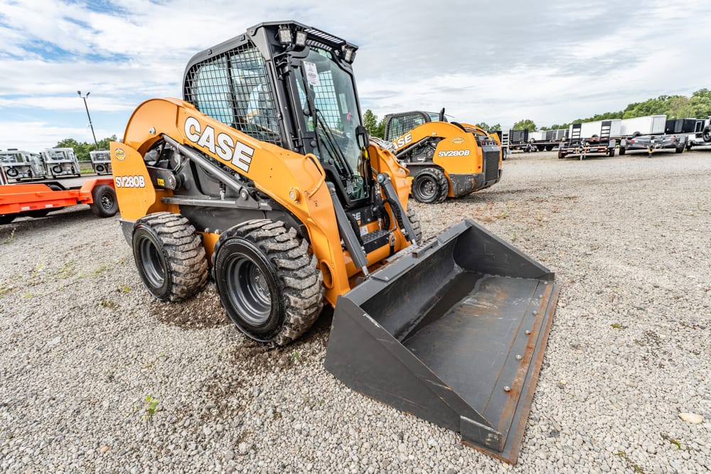 Front view of an orange CASE skid-steer loader with a large bucket, parked on a gravel lot with other heavy equipment in the background.