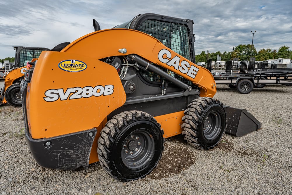 Orange CASE SV208B skid-steer loader on a gravel lot with a front bucket and other equipment in the background