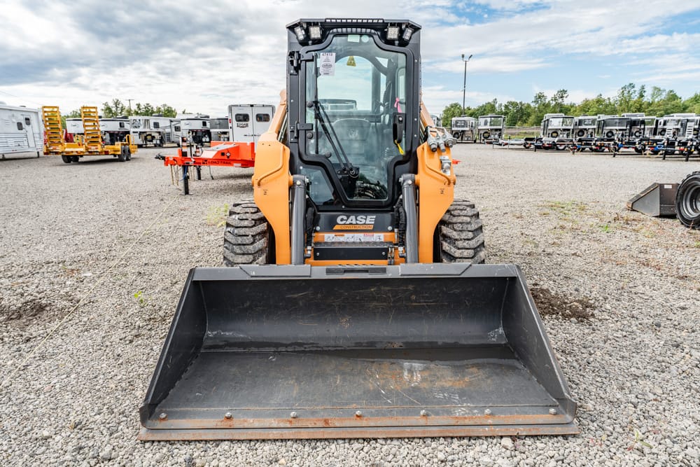 Orange CASE skid-steer loader with a wide front bucket facing the camera in a gravel lot, with trailers and equipment in the background.