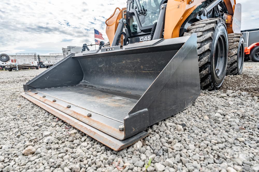 A close-up of a heavy construction skid steer with a large metal bucket attachment resting on rocky ground, with an American flag and trailers in the background.