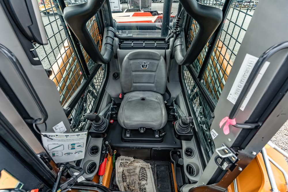Operator sits in the cockpit of a construction excavator, with dual joysticks, protective bars, and a view to the outside yard.
