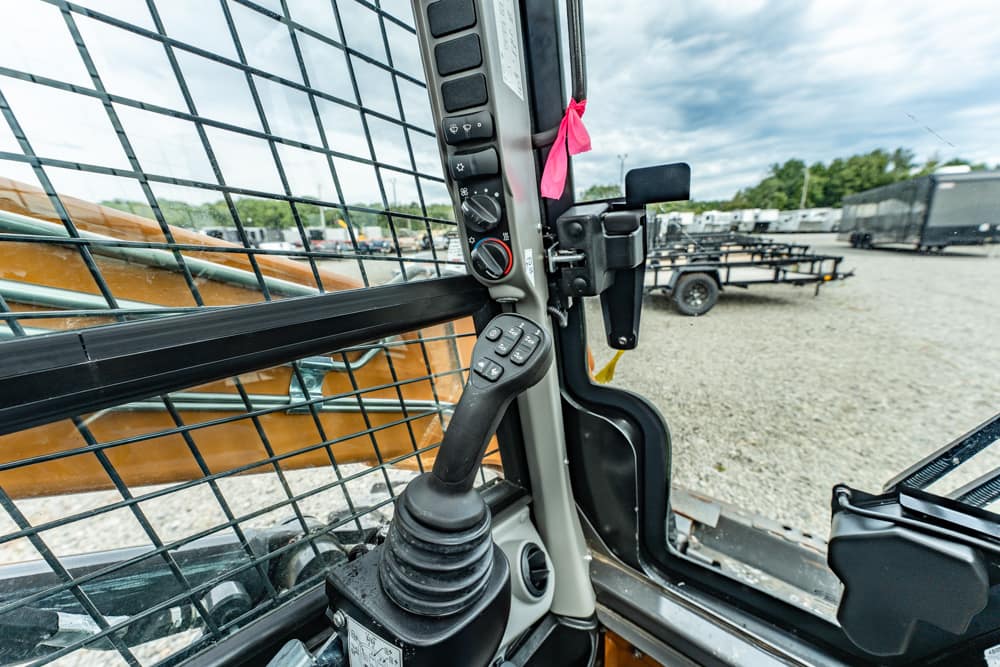 Close-up of a joystick and control panel inside a construction vehicle cab, with a metal mesh window and trailer yard visible outside.