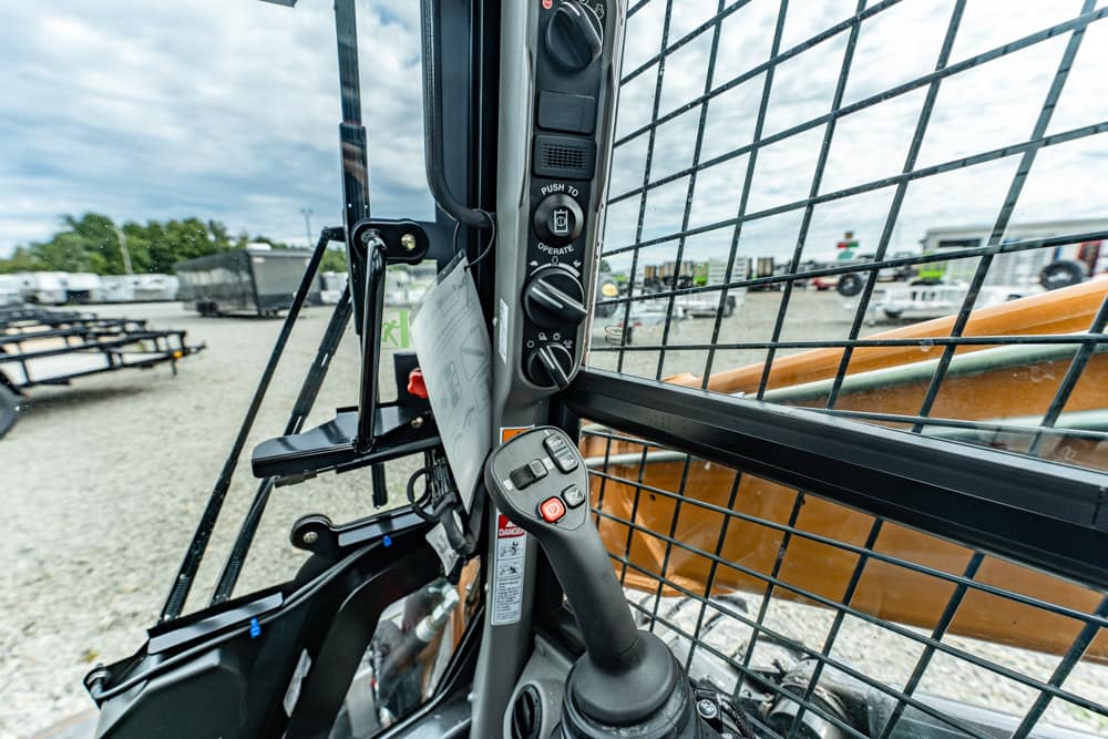 Inside the cab of a construction skid-steer loader, showing the joystick, control panel, and protective mesh while parked in a yard.