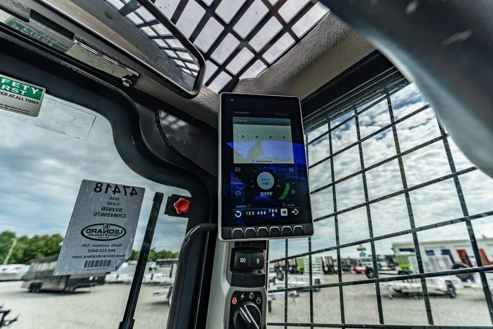Cab interior of a heavy equipment cockpit with a mounted touchscreen tablet showing controls, viewed through an open door with a metal grille and a yard outside.