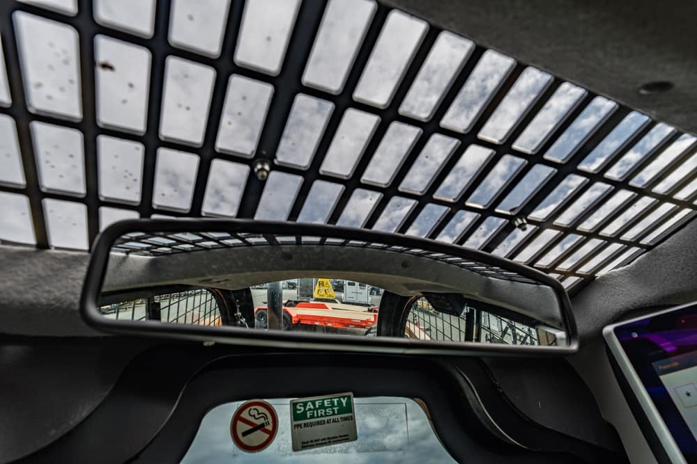 View from inside a vehicle: rearview mirror shows a construction yard with orange equipment beyond a fence; metal grid overhead and safety stickers nearby.