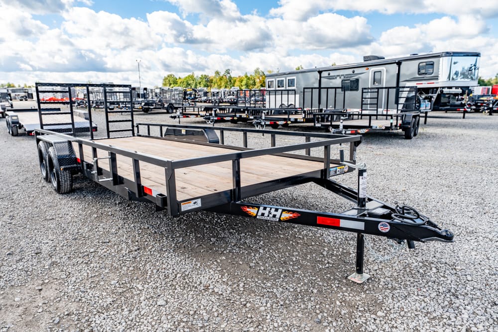 Black dual-axle flatbed trailer with a wooden deck on a gravel lot, surrounded by other trailers and an RV in the background.