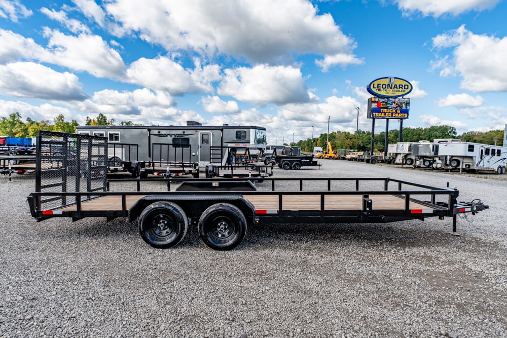 Wide flatbed trailer with wooden bed and black metal rails in a dealership lot, several trailers in the background.