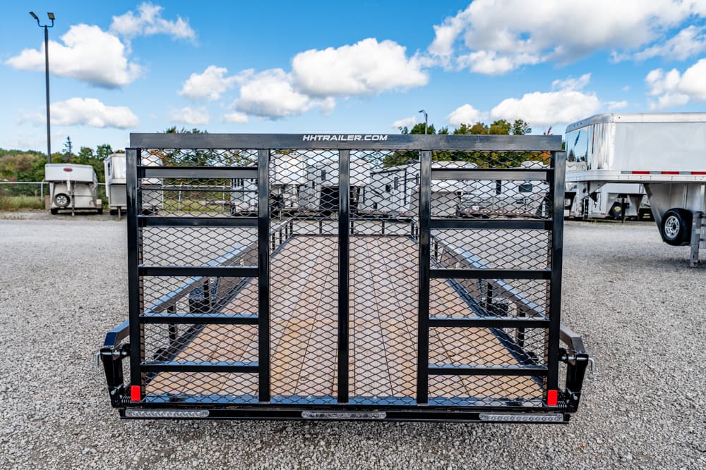 Black utility trailer with a mesh ramp gate, wooden bed, and dual side rails in a gravel lot under a blue sky.