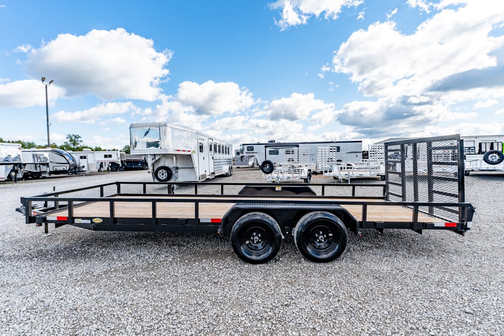 Dual-axle flatbed trailer with a wooden deck and a mesh rear gate, parked on a gravel lot with RVs in the background.