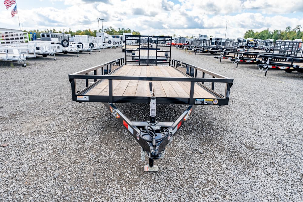 Front view of a flatbed utility trailer with a wood deck and metal rails in a gravel trailer yard, surrounded by other trailers.