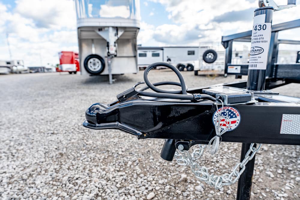 Close-up of a black trailer hitch with a coiled cable and safety chains, in a gravel lot with other trailers in the background