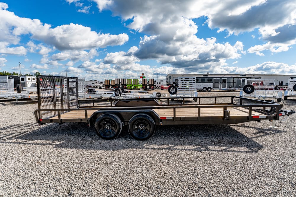 Flatbed utility trailer with dual axles on a gravel lot, with other trailers and buildings in the background under a blue sky with clouds.
