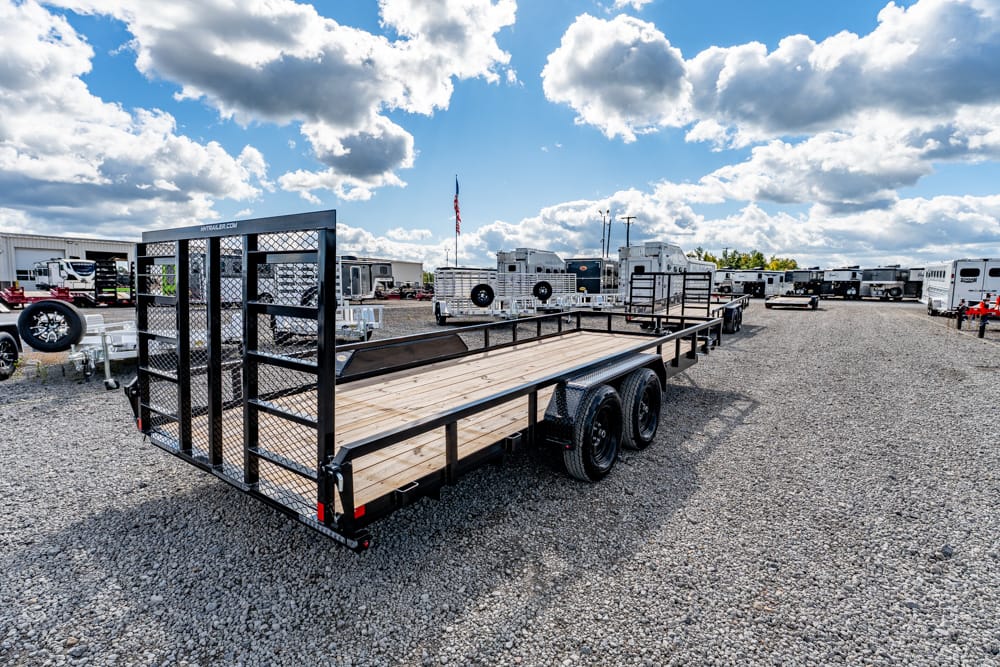 Flatbed utility trailer with a ramp gate parked on a gravel lot among other trailers under a blue, cloudy sky.