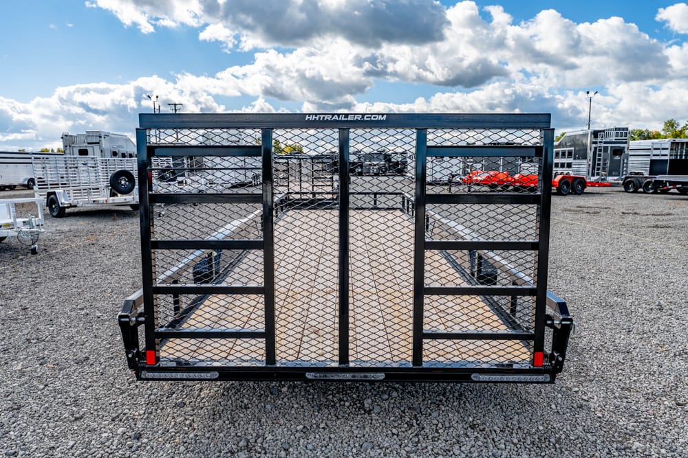 Rear view of a black utility trailer with a diamond-m mesh gate on a gravel lot, with other trailers in the background at a dealership center.