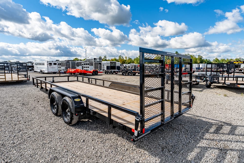 Flatbed utility trailer with a rear mesh gate, parked on a gravel lot among other trailers and equipment