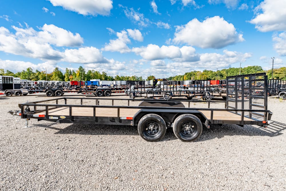 Flatbed trailer with wooden deck and railing parked on a gravel lot, with other trailers in the background under a blue sky.