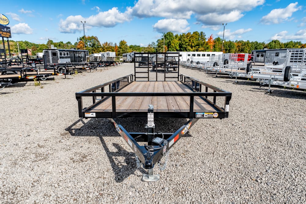 Front view of a black utility trailer with a wooden deck on a gravel lot, surrounded by rows of trailers in a dealership yard.