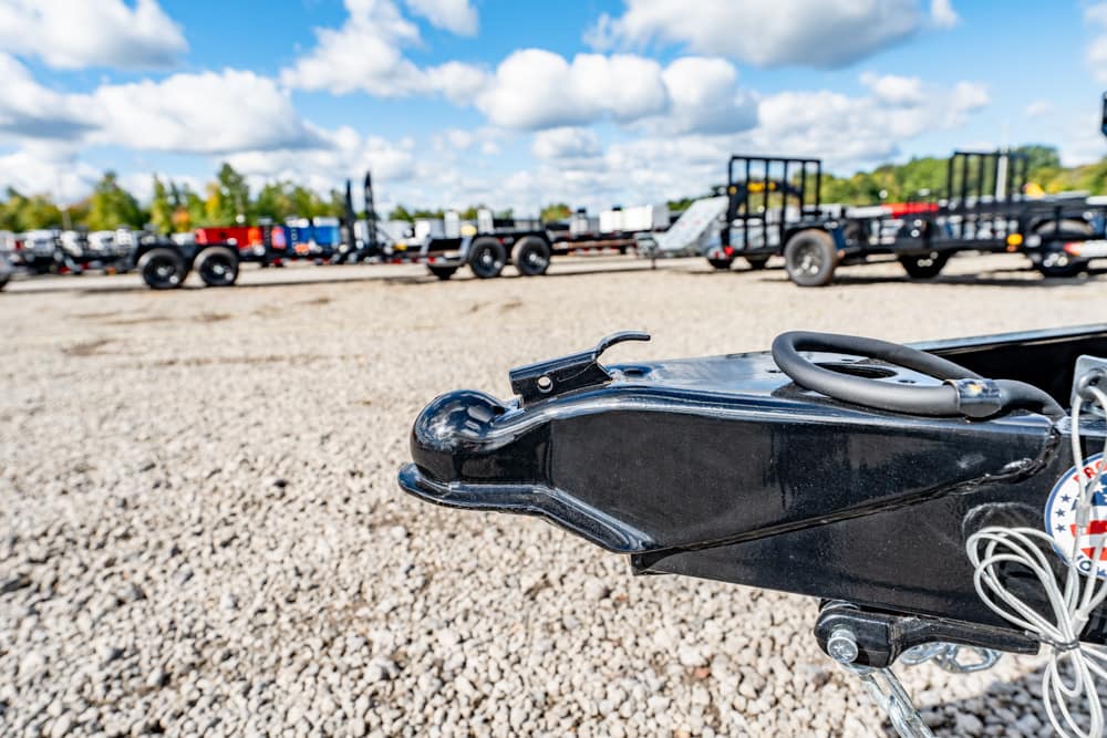 Close-up of a black trailer hitch/coupler on a gravel lot with other trailers in the background.