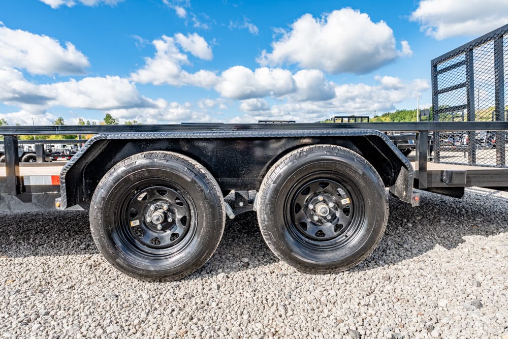 Dual-axle utility trailer with black metal fenders on a gravel lot under a bright, partly cloudy blue sky