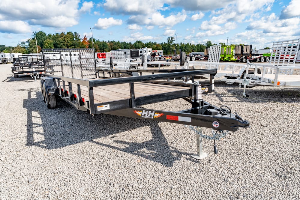 Black flatbed trailer with wooden deck and metal rails on a gravel lot; other trailers and equipment visible in the background.