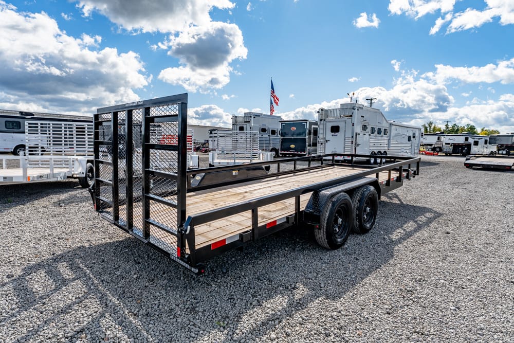 Flatbed trailer with a wooden deck and metal gate parked on a gravel lot, other trailers and RVs in the background.