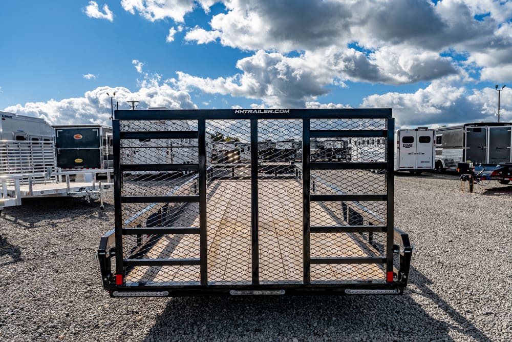 Front view of a black utility trailer with a mesh ramp gate, parked on a gravel lot among other trailers; clear blue sky above.