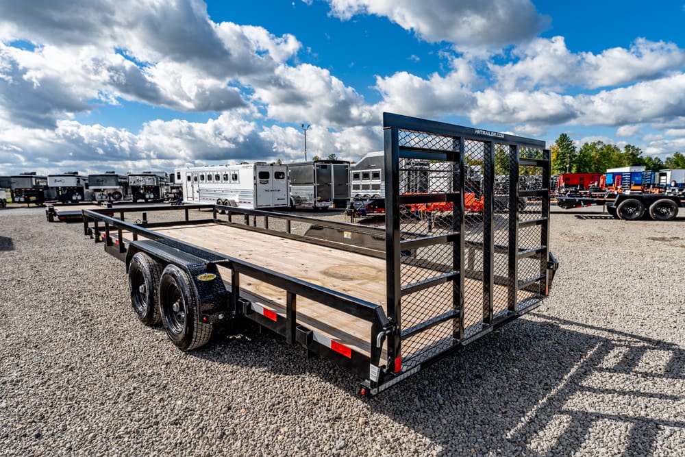Dual-axle black flatbed trailer with a tall mesh gate, parked on a gravel lot among other trailers.